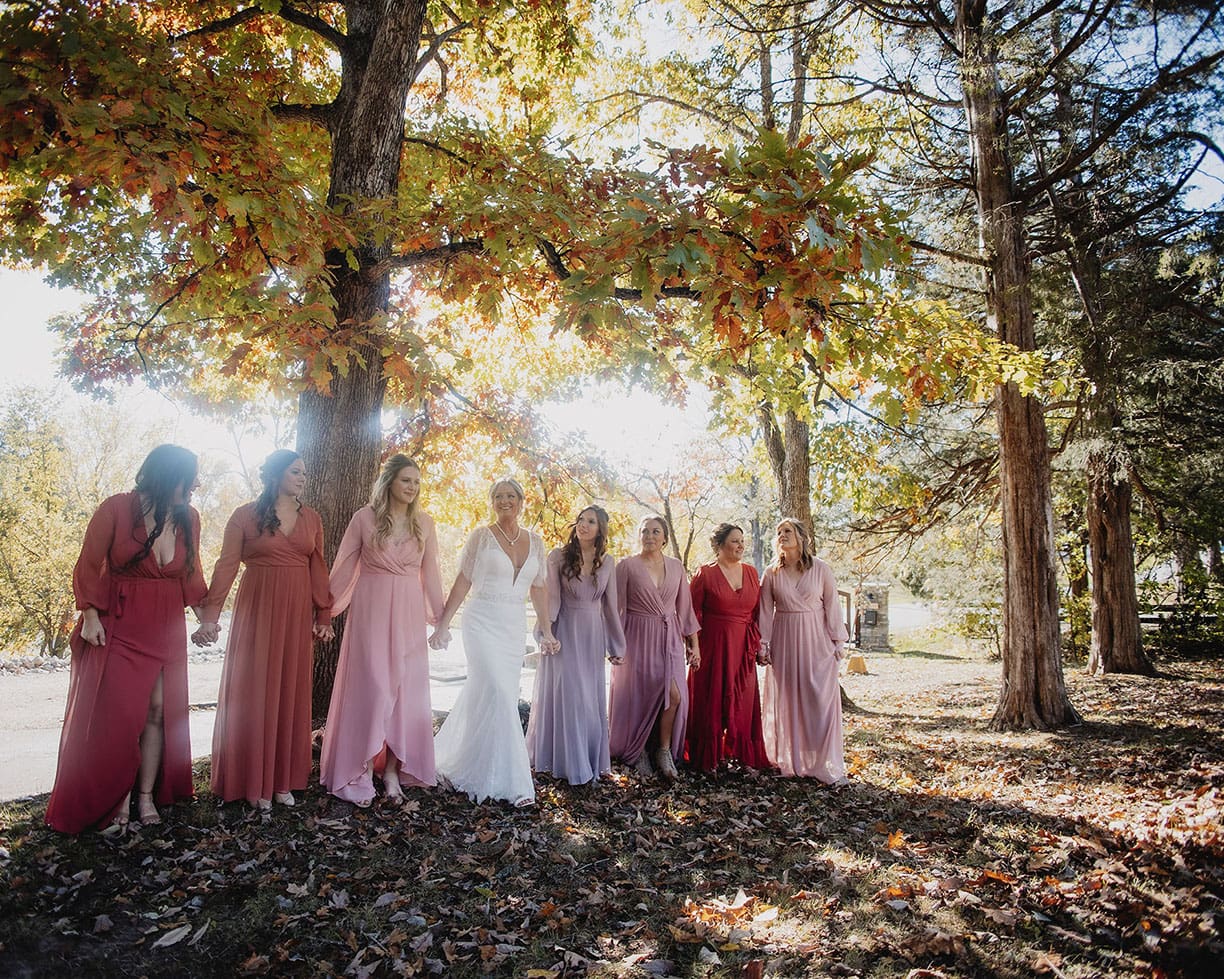 Bride and bridesmaids walking hand-in-hand through the fall leaves with the sun illuminating behind them.