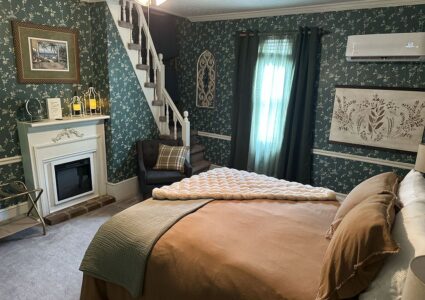This is an image of the Bridal Suite at Ross Mansion at The Barns at Marengo Cave. It shows a queensize bed with nice covers and a luxuriously soft cream colored throw blanket. There is a chair, artwork and a remote controlled fireplace in the room.