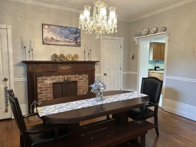 This is the dinig room at Ross Mansion at The Barns at Marengo Cave. Image shows a long narrow dining table with blue flowered runner and a vase of flowers in the middle. A beautiful period chandelier hangs above the table and a fireplace is located in the background.